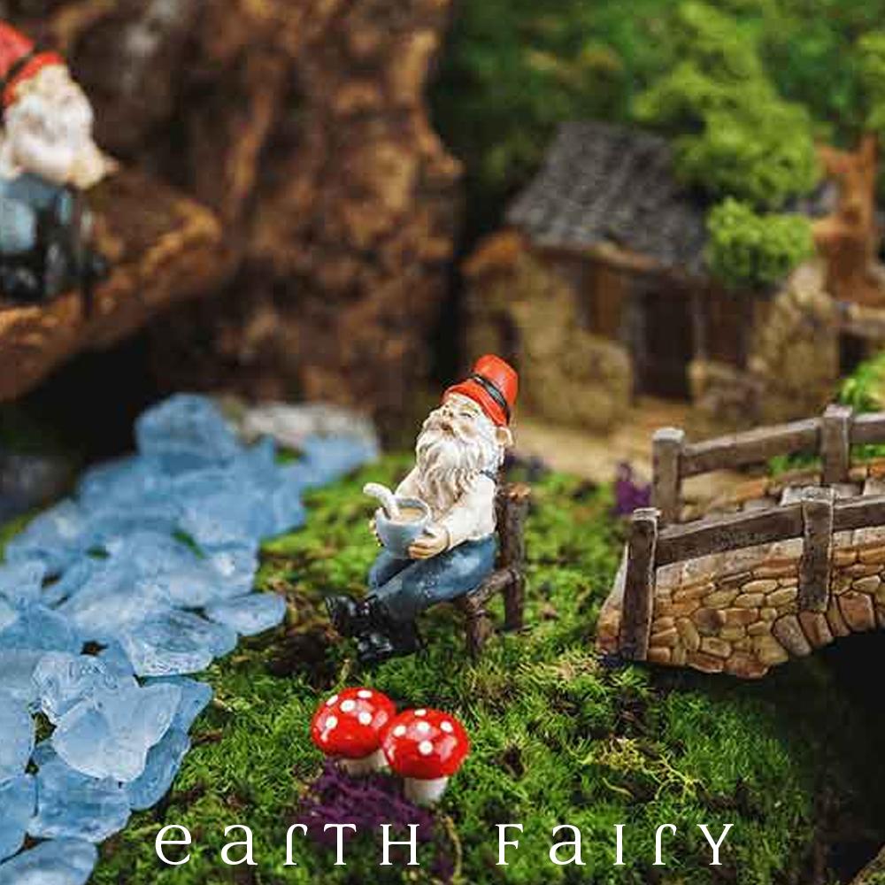 miniature polyresin gnome figurine, posed sitting on a log hewn chair, black boots, blue pants, cream top and red pointy hat, holding a mug of coffee