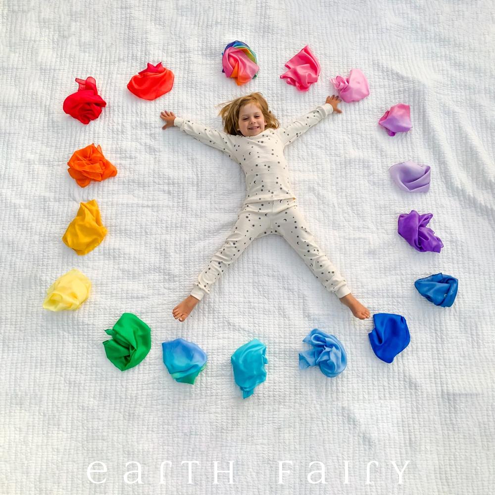 Mini Playsilks, showing child surrounded by rainbow of playsilks in various colours, from The Earth Fairy Silk Collection