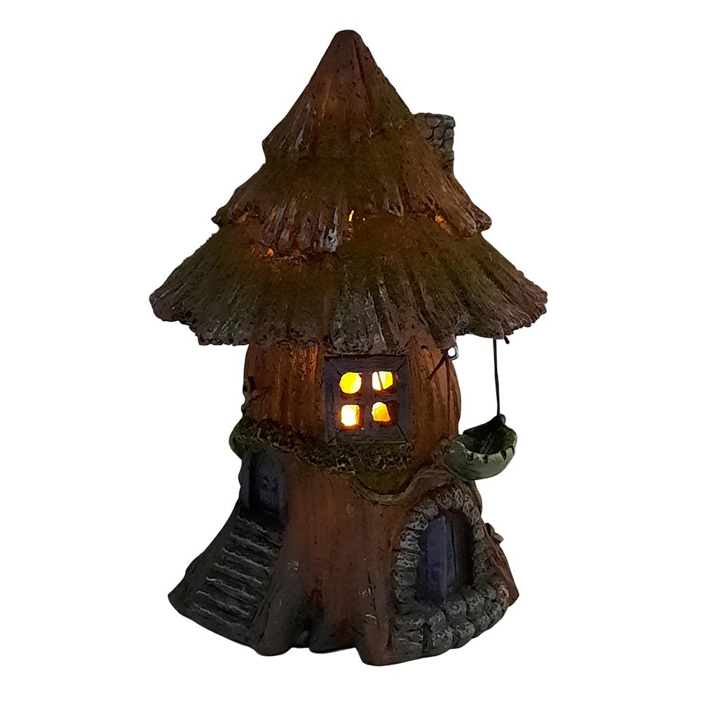 Night time view with lights on of the Moss Tree House with Hammock - Solar, a miniature fairy house for the garden with mossy wooden roof and purple arched door, and a small hammock hanging to the side