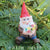miniature polyresin gnome figurine, wearing boots, blue pants, orange coat and red pointy hat, posed sitting and clasping a bunch of bright flowers
