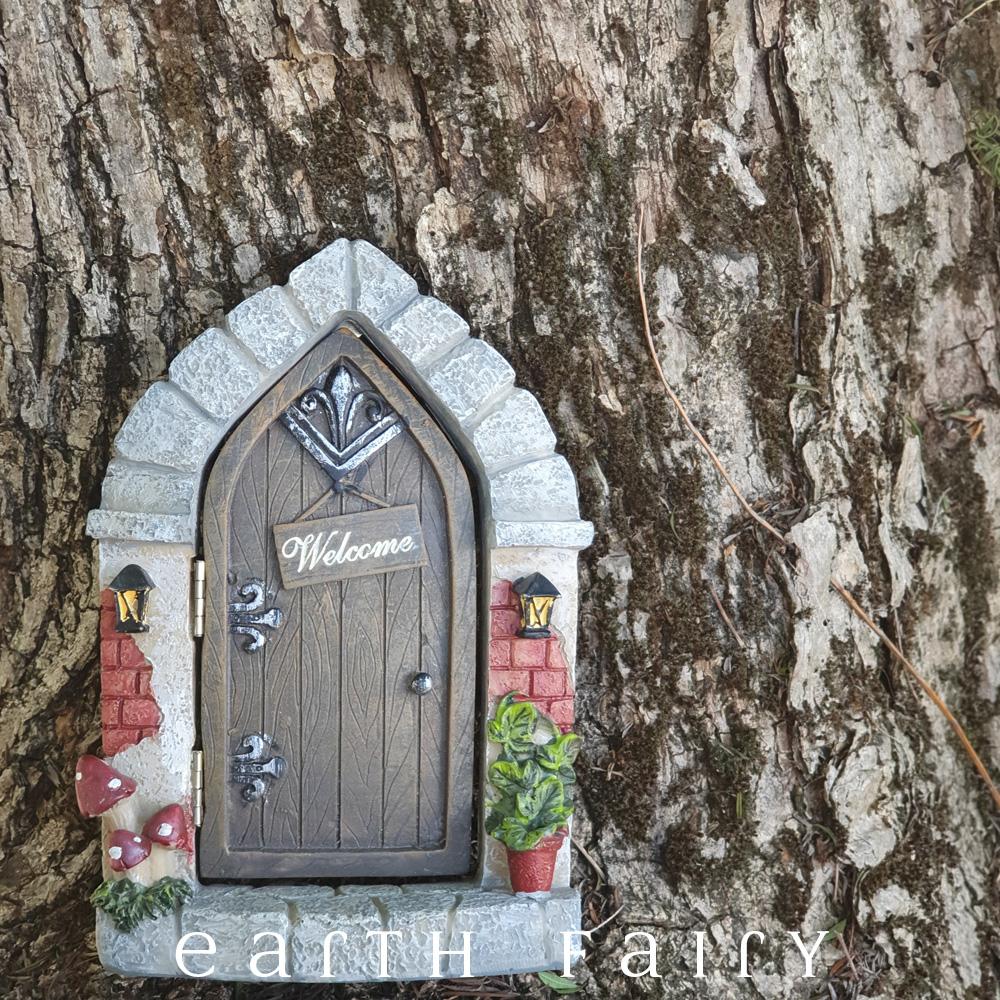 polyresin miniature fairy door, arched cobblestone frame, with wood effect door and hanging "welcome" plaque, toadstools to one side, pot plant on the other side